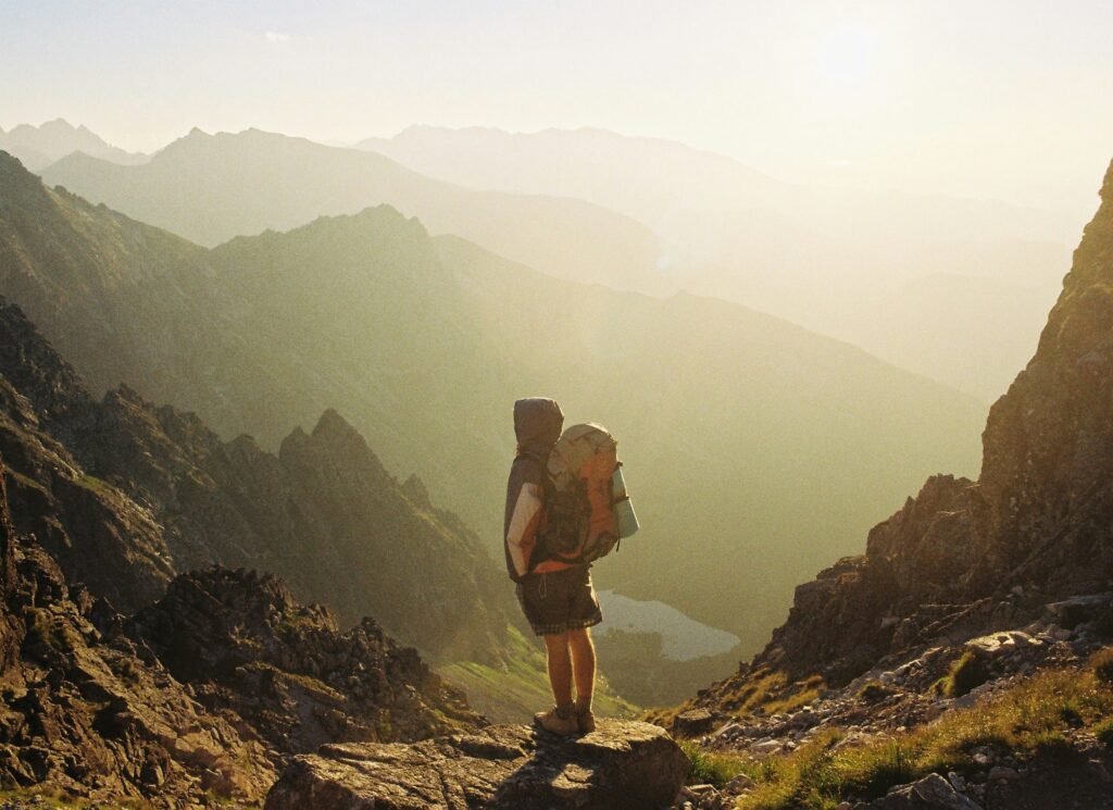 7 Eine alleinstehende Wanderin mit großem Rucksack blickt von einem felsigen Bergpass in ein sonnenbeschienenes Tal mit sanften Hügeln und einem See in der Ferne.
