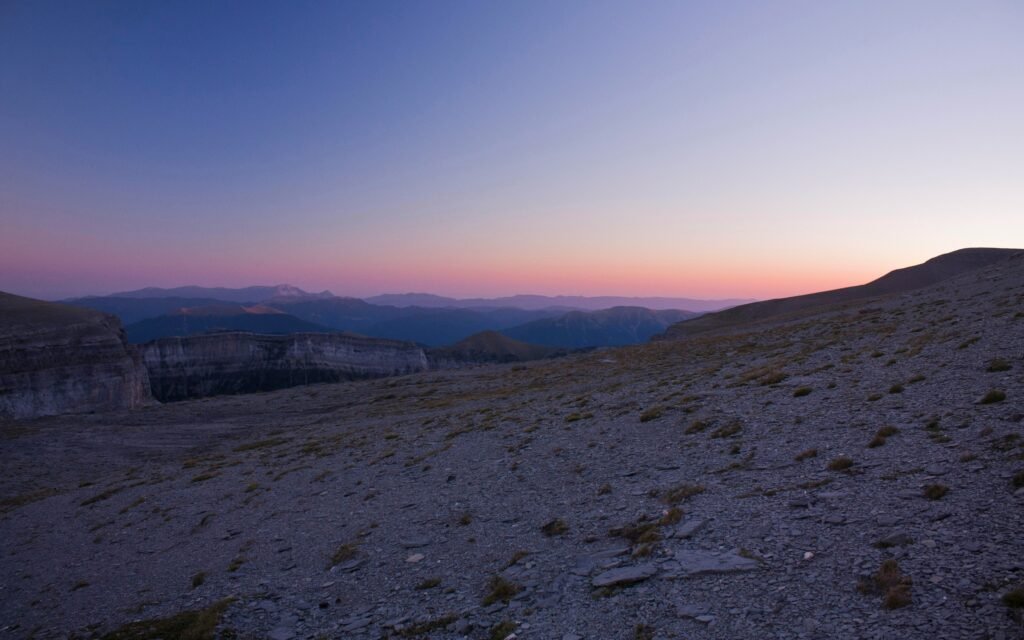 9 Weite, steinige Berglandschaft im sanften Licht der Dämmerung, mit violett-orangem Himmel über einem entfernten Horizont aus Bergketten.